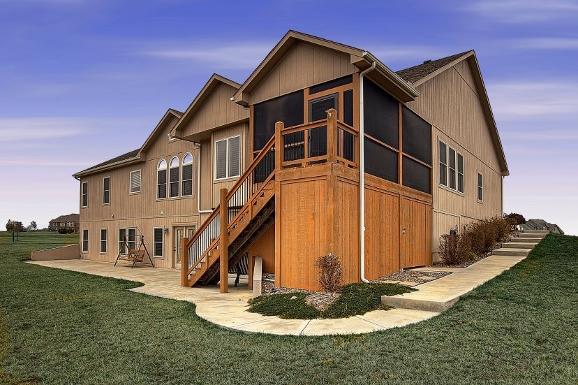 A tan house with a wooden screened-in porch, stairs leading to a stone patio, and a grass yard under a blue sky.