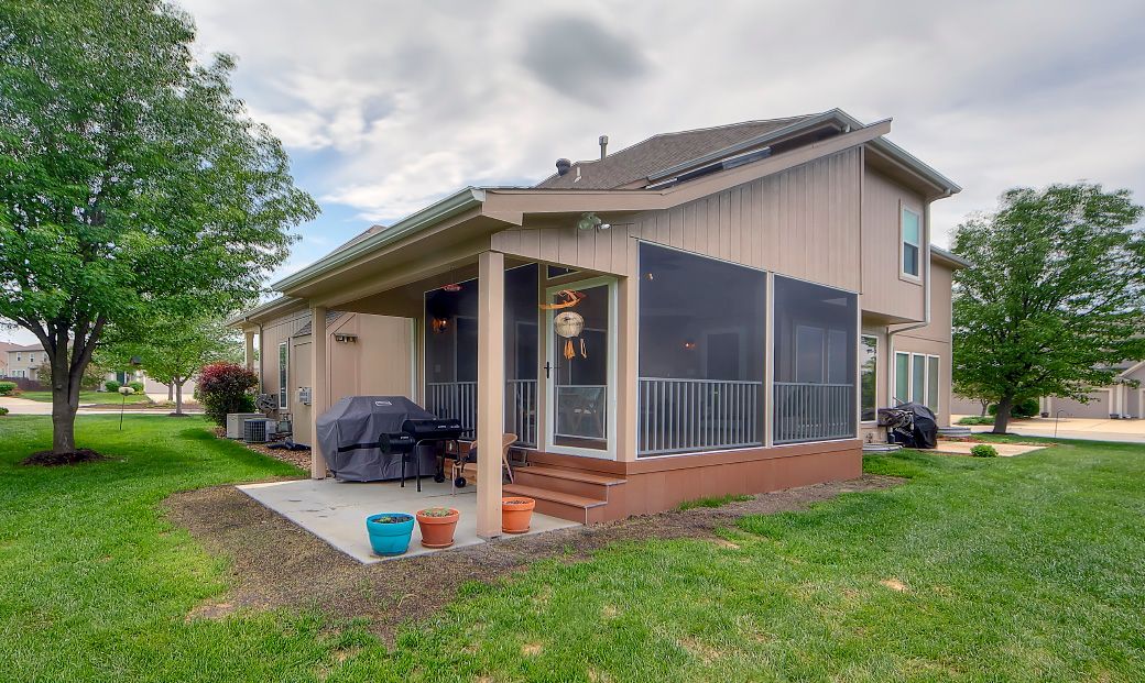 A tan house with a screened-in porch and a concrete patio in a grassy backyard with trees on a cloudy day.