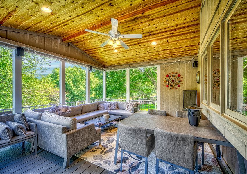 A screened-in porch with wood plank ceiling, light gray sectional sofa, dining table with chairs, and lush green trees.