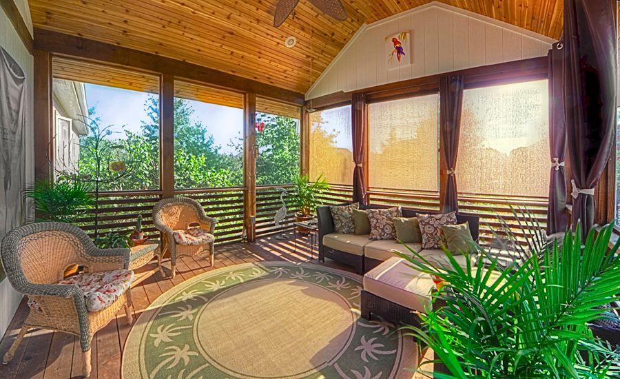 A screened-in porch with wicker chairs, a sofa, a round area rug, and lush greenery under a wooden ceiling.