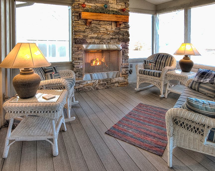 A sunroom features a stone fireplace with a fire, white wicker chairs with patterned cushions, and two table lamps.