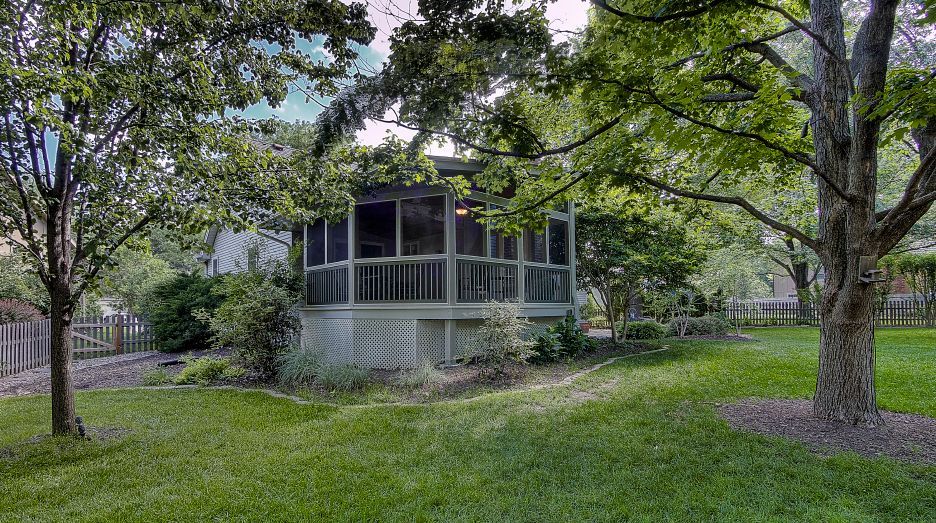 A screened-in porch with light-colored siding sits nestled in a grassy backyard, surrounded by mature, leafy green trees.