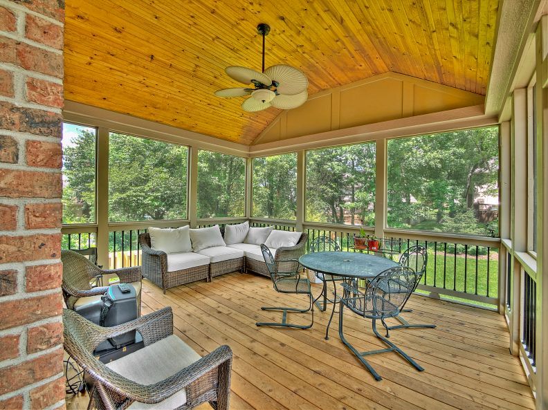 A screened-in porch with a wooden ceiling, wicker furniture, and a round metal table, overlooking a backyard.