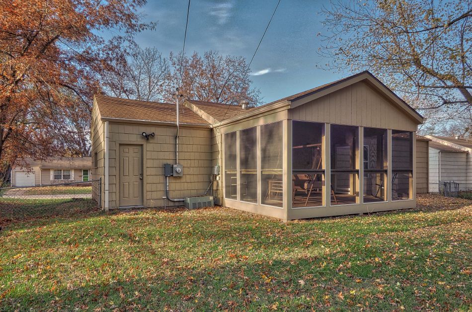 A beige house with a sunroom and brown roof, surrounded by fall foliage in a yard covered with fallen leaves.