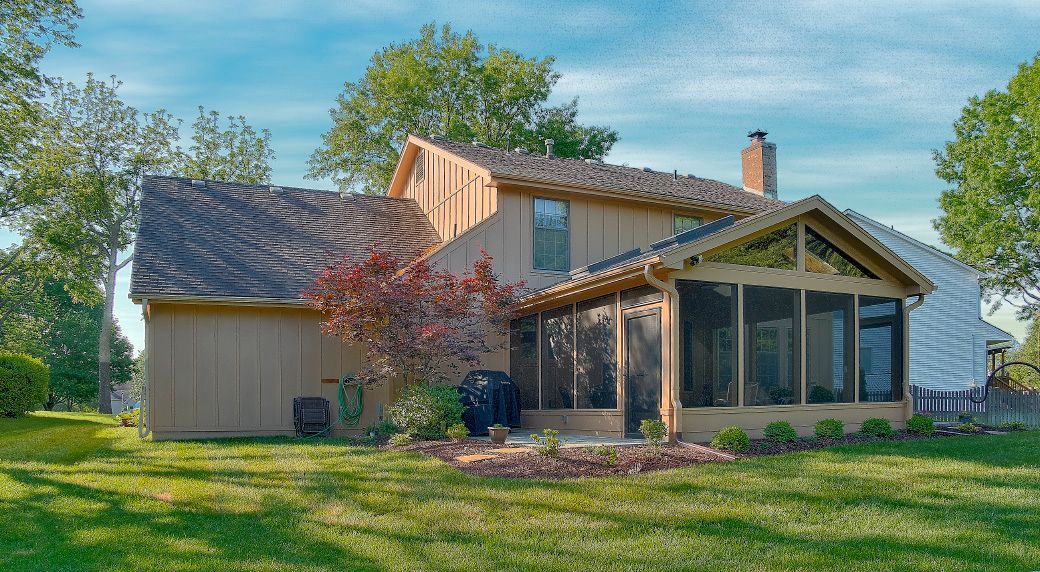 A tan, two-story house with a large screened-in porch on the back, situated on a green lawn with trees under a blue sky.