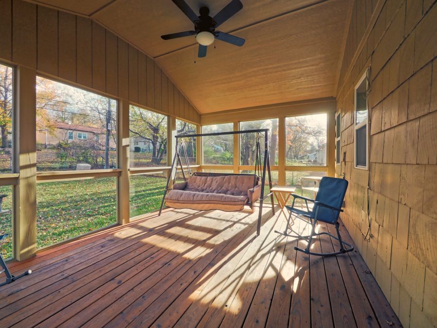 A wooden screened-in porch featuring a hanging swing bench, a rocking chair, and a ceiling fan overlooking a grassy yard.
