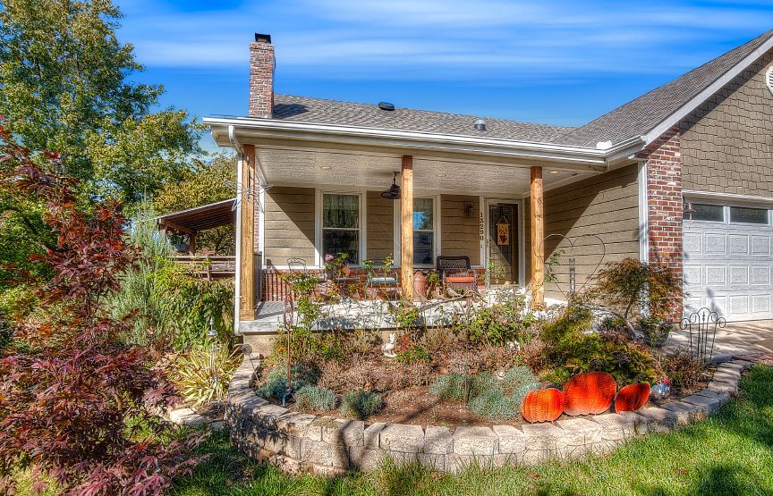 A tan brick house features a covered front porch, stone landscaping, and large orange pumpkins on a sunny day.