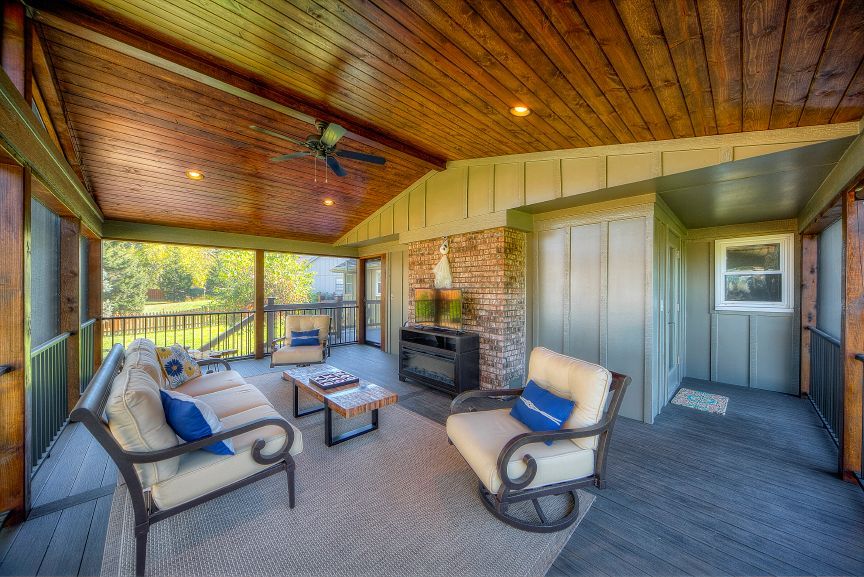 A screened-in porch with a wood-plank ceiling, stone fireplace, couch, armchair, and views of a grassy yard.