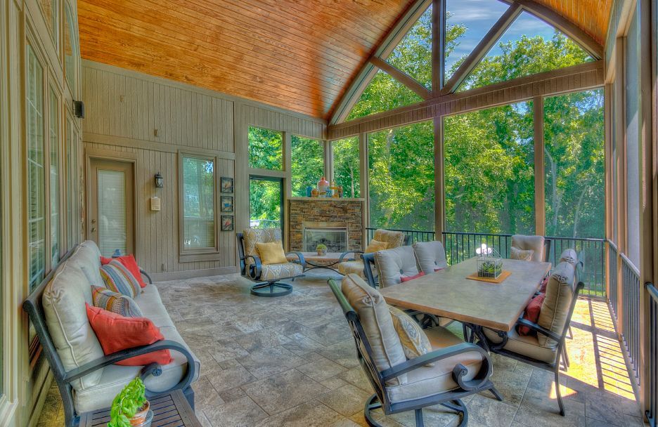 A spacious screened-in porch with a wooden vaulted ceiling, stone fireplace, dining table, and seating overlooking trees.