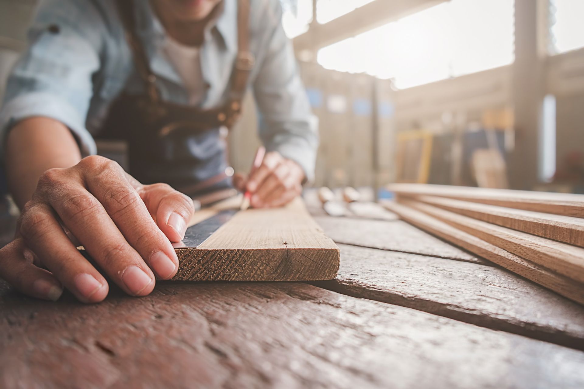 A craftsperson uses a pencil and metal ruler to measure a wooden plank on a workbench in a bright workshop.