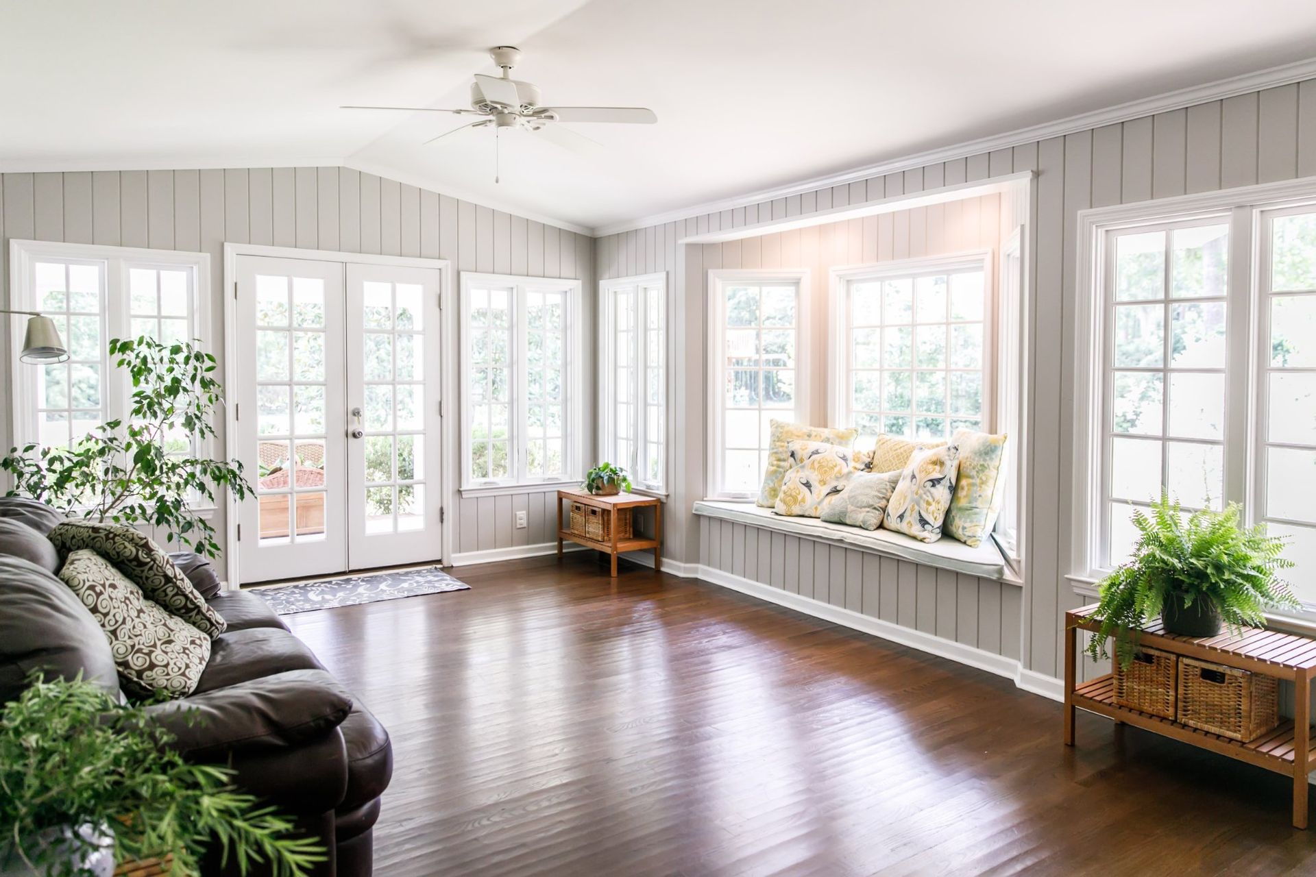 Bright living room with white paneled walls, dark wood floors, a cozy window seat, potted plants, and French doors.