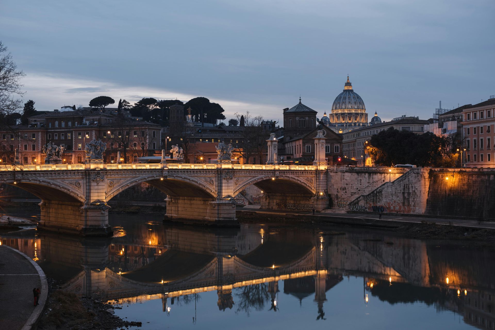 A bridge over a river in a city at night with a dome in the background.