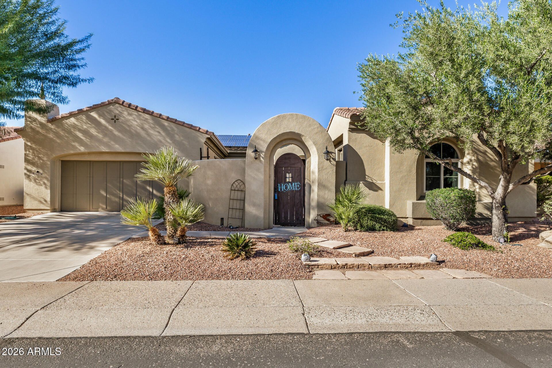 Single-story stucco house with a two-car garage, arched entry, and desert landscaping under a blue sky