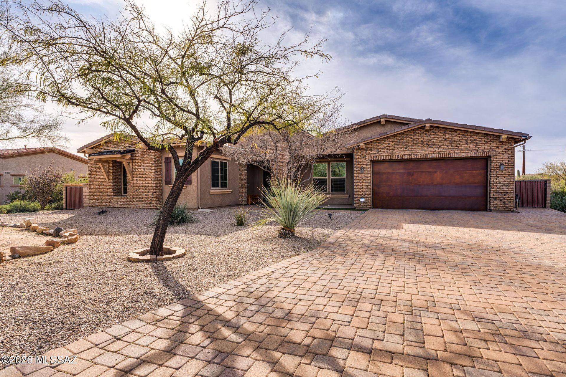 Desert-style house with stone facade, brick driveway, and a tree in the front yard under a blue sky