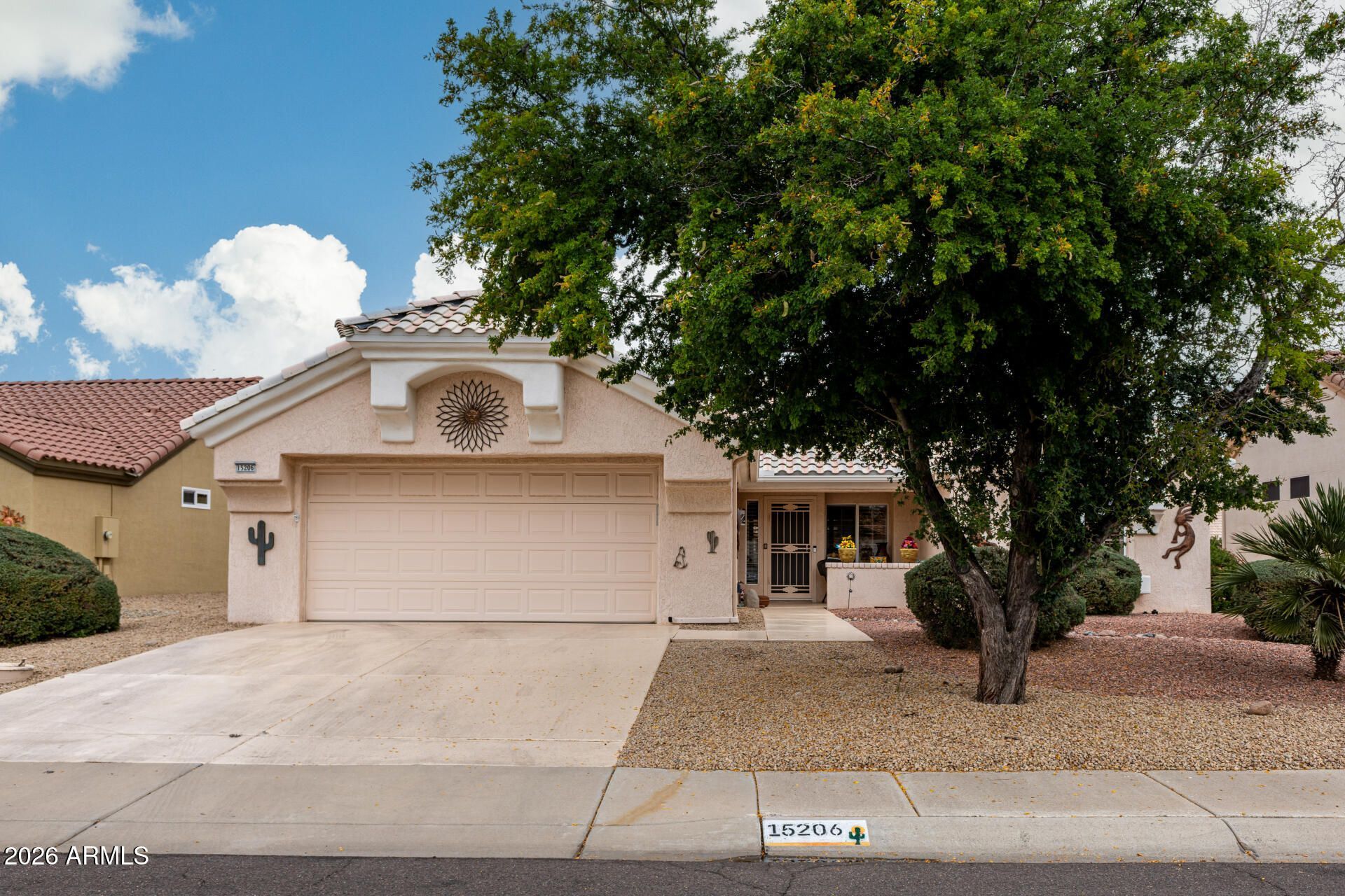 Single-story beige suburban house with a two-car garage, tree, and desert landscaping under a partly cloudy sky