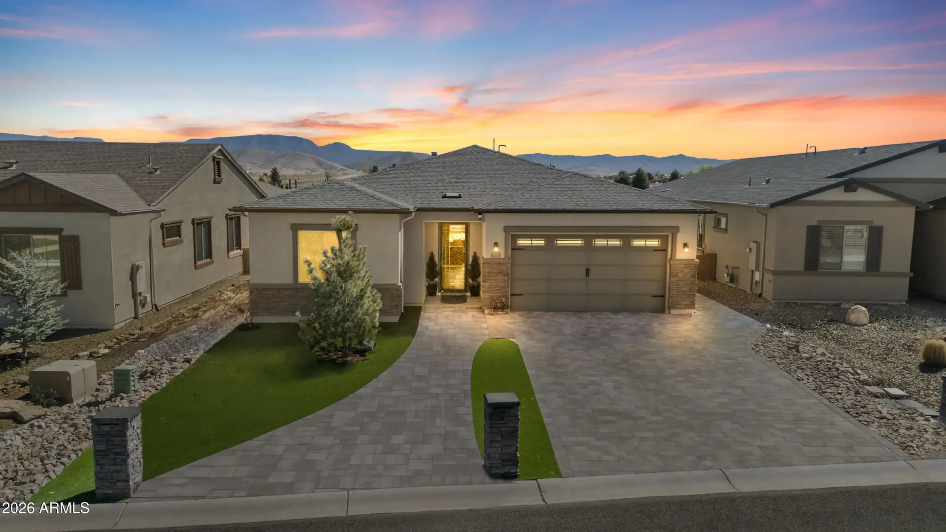 Modern single-story house at sunset with a lit entryway, two-car garage, and stone driveway.