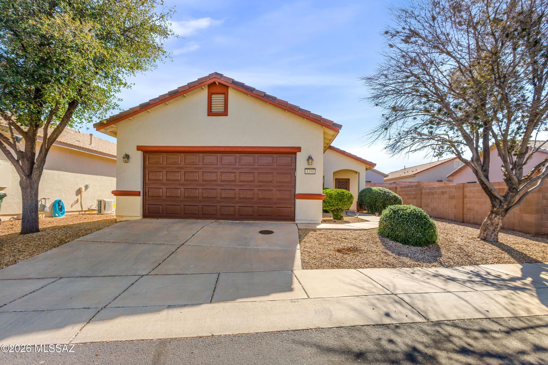 Single-story beige house with two-car garage, desert landscaping, and a driveway in front.