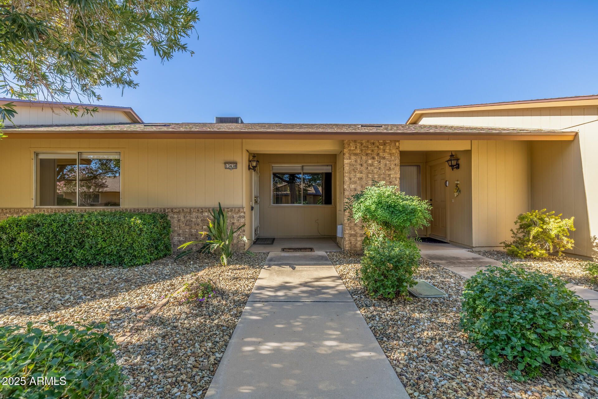 Single-story beige house with a front walkway, porch, and desert landscaping under a clear blue sky