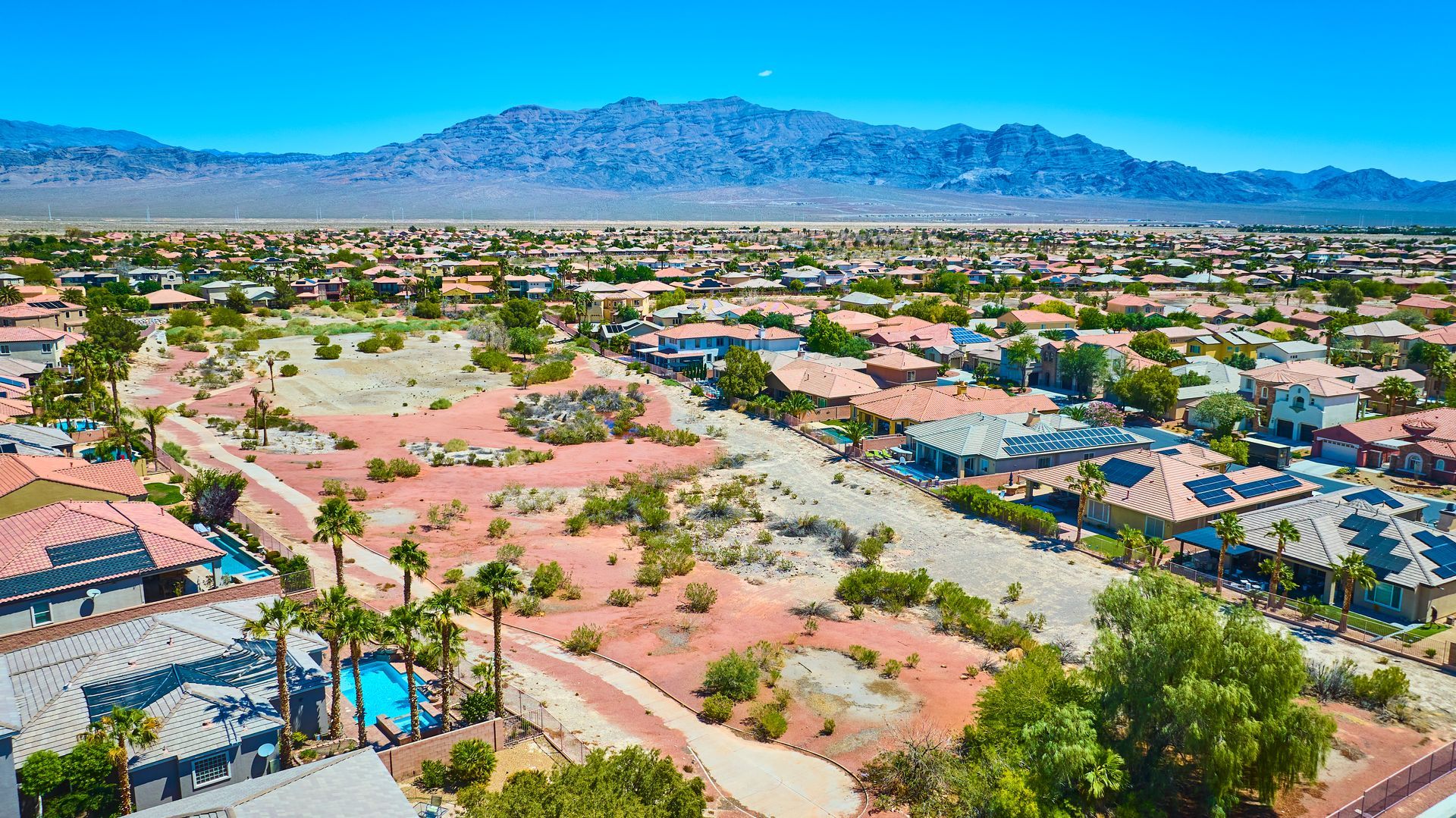 Aerial view of a desert suburb with houses, palm trees, and mountains under a clear blue sky