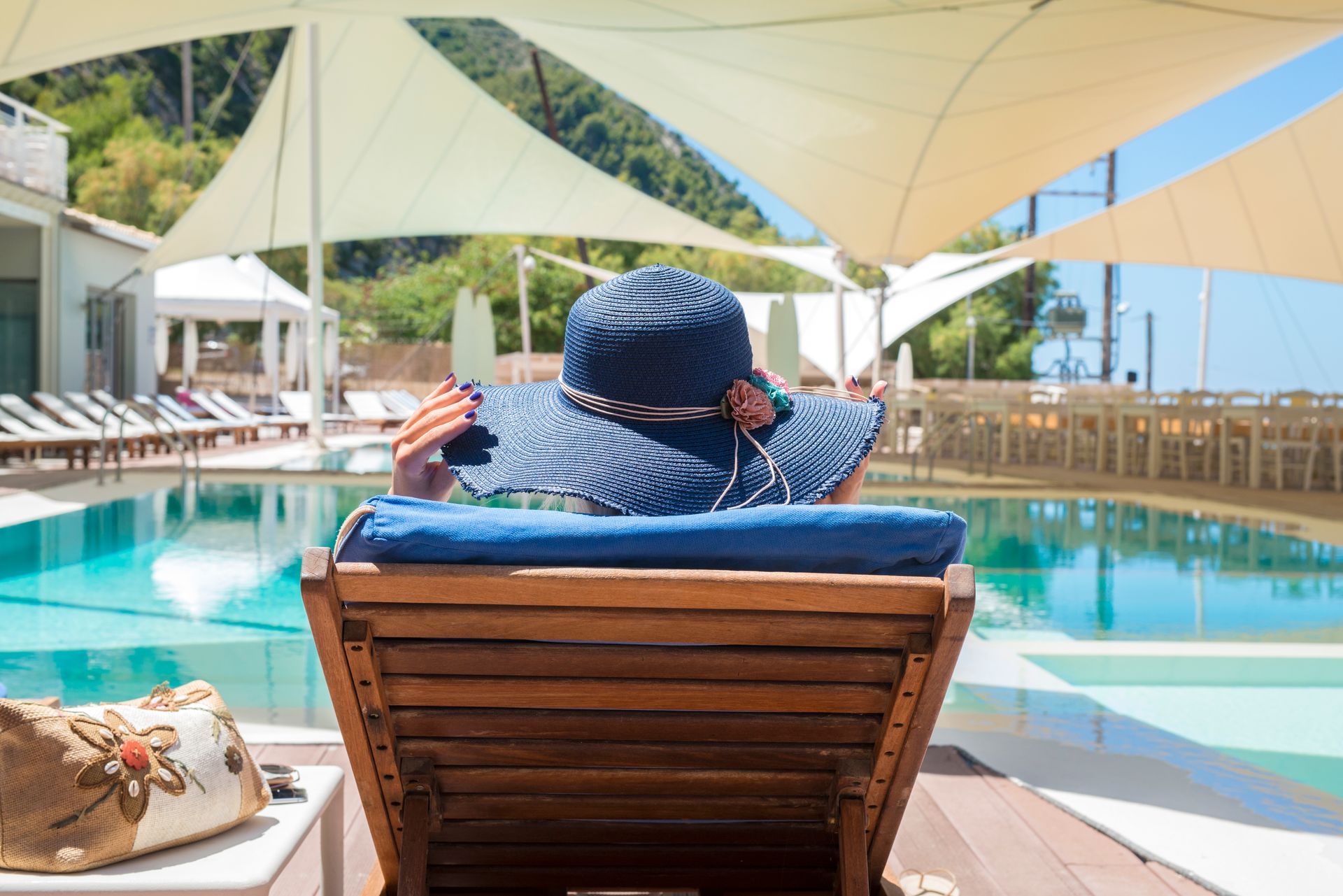 Person relaxing on a lounge chair by a sunny resort pool under white shade sails