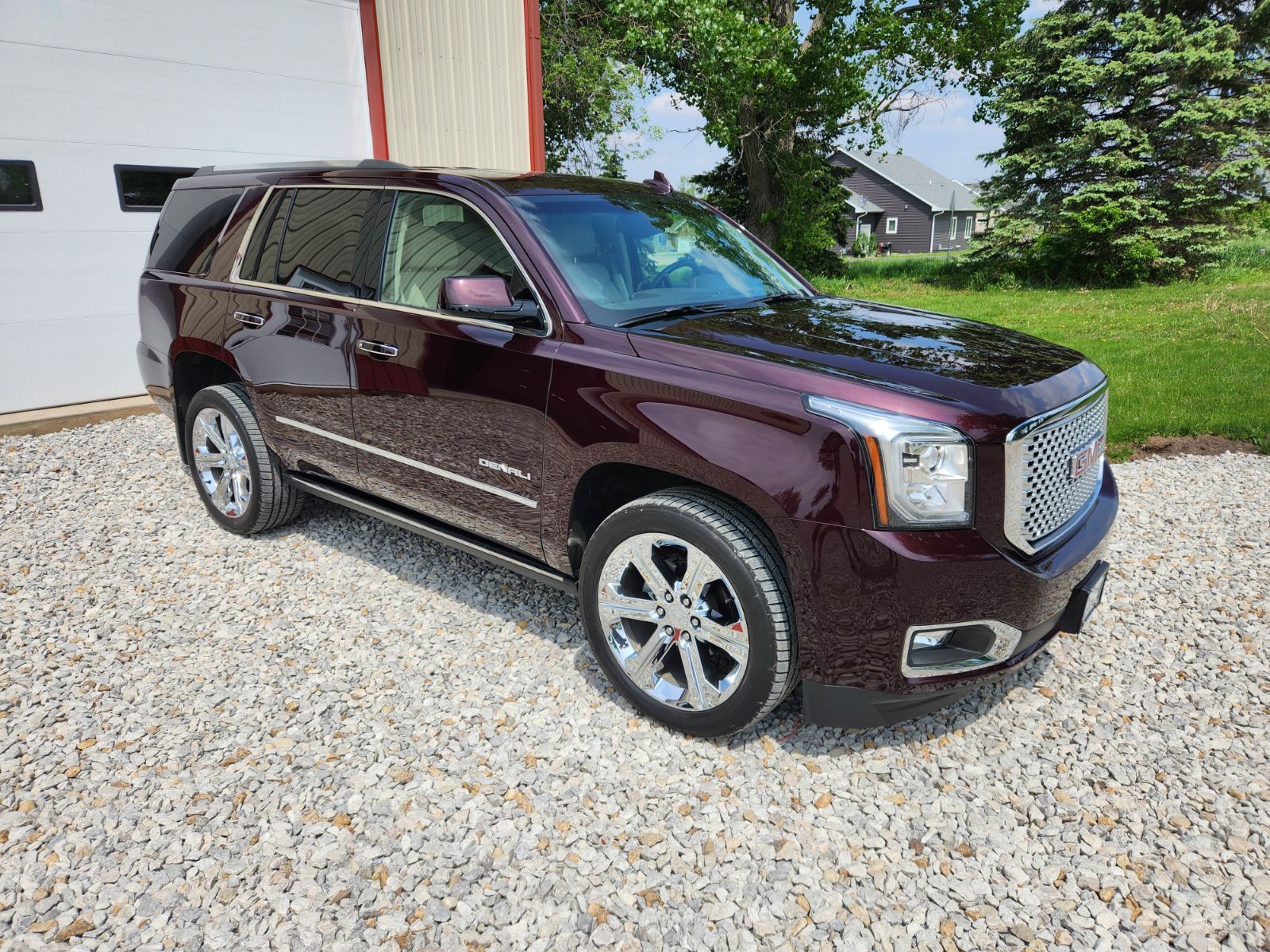 A burgundy gmc yukon is parked in a gravel lot in front of a garage.