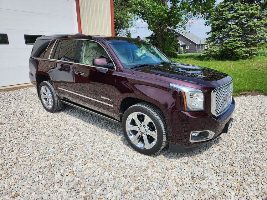 A burgundy gmc yukon is parked in a gravel lot in front of a garage.