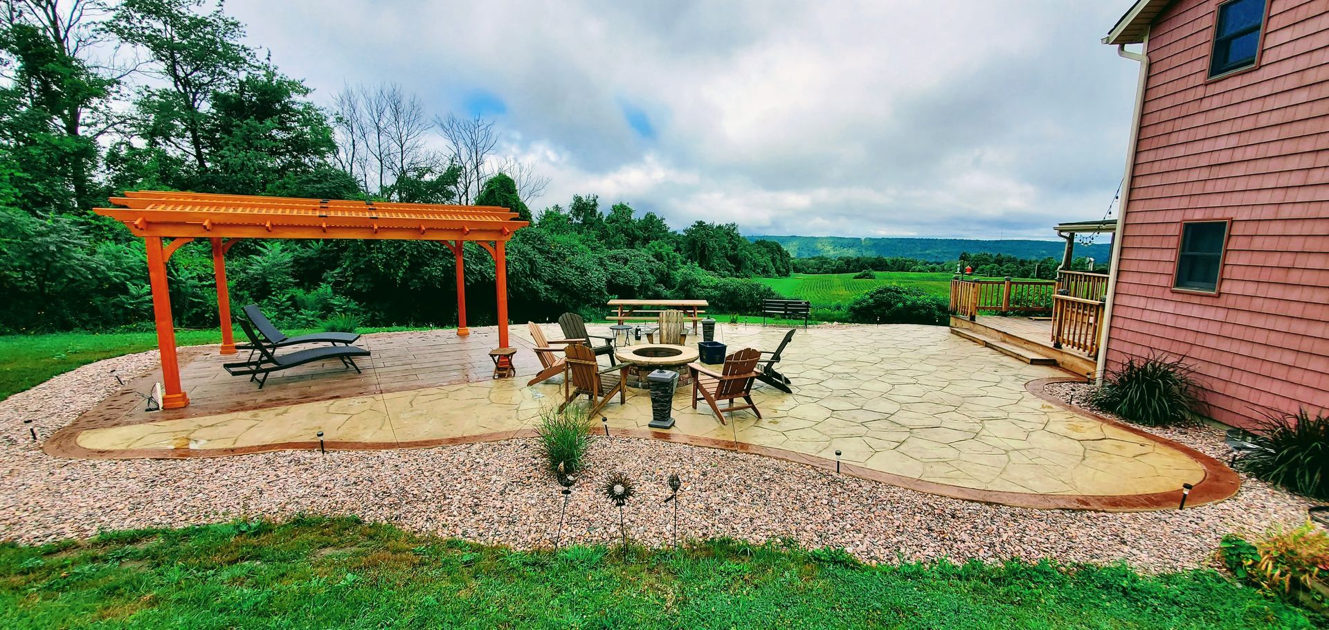 A wooden pergola and chairs on a patio, with a pink house to the right and green landscape in the background.