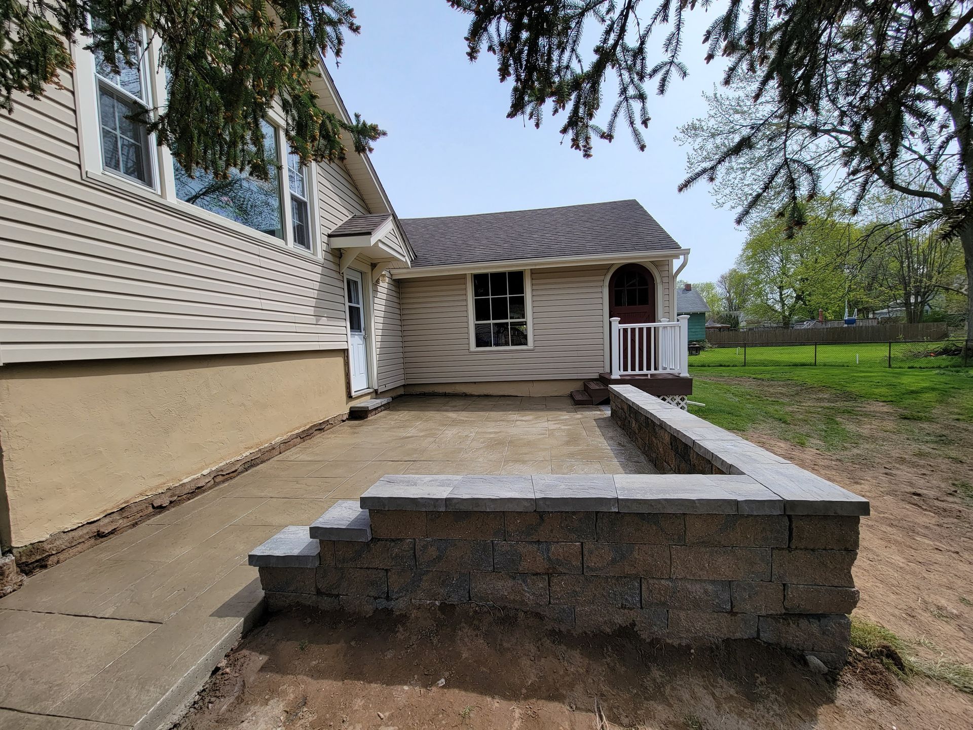 Exterior view of a house with a new retaining wall, patio, and small entrance on a sunny day.
