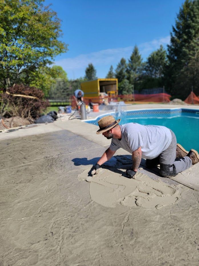Man kneels, smoothing cement near a pool. He wears a hat and gloves. Sunny day.