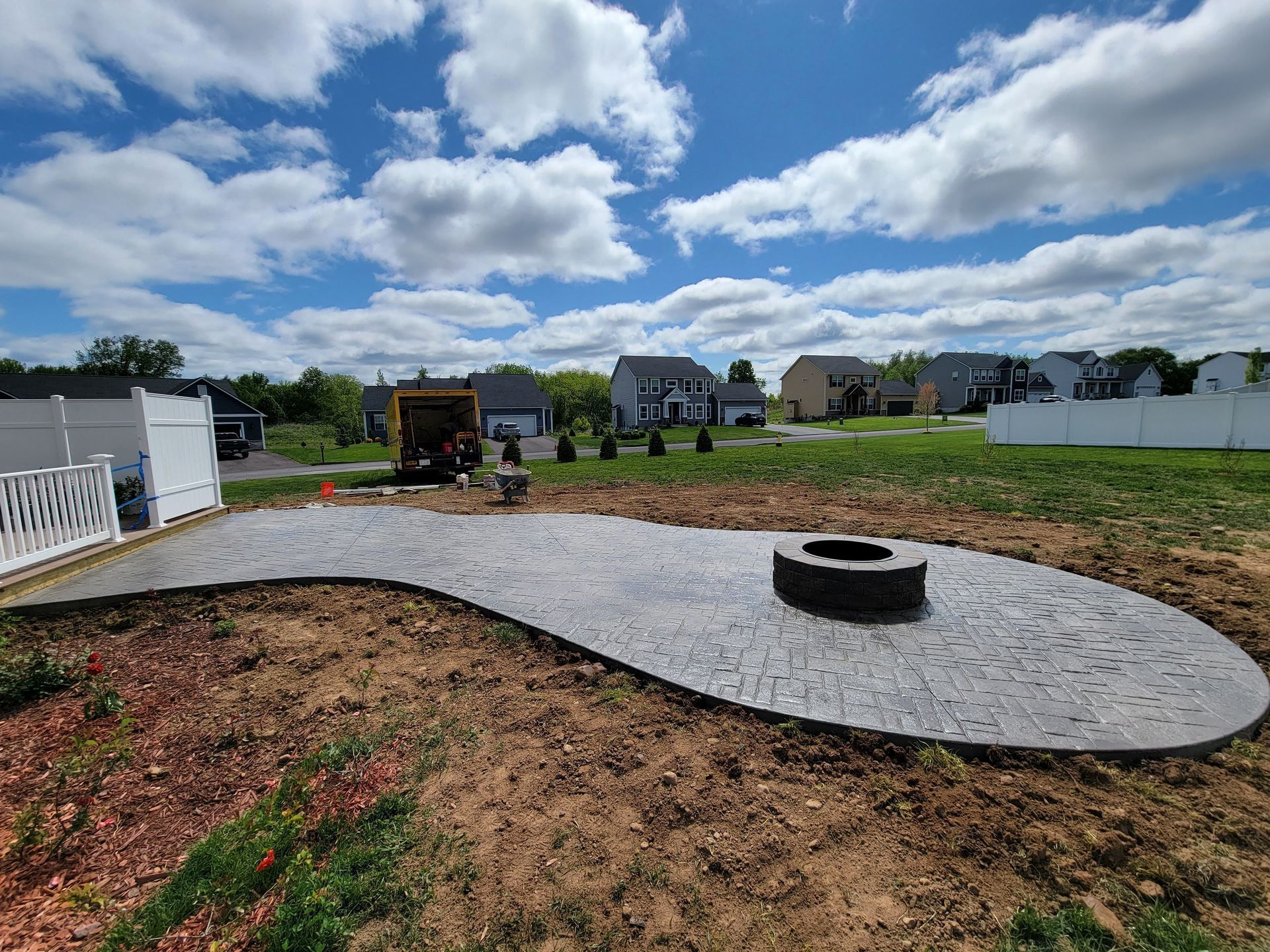 A newly constructed gray stone patio with a fire pit in a suburban backyard on a sunny day.