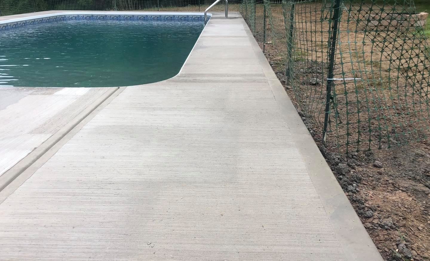 Concrete pool deck next to a turquoise pool and a chain-link fence on a sunny day.