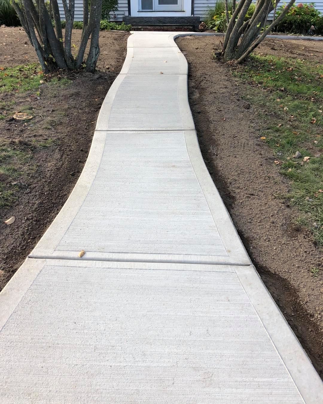 Concrete walkway leading to a house. Light gray concrete with soil and shrubs on either side.