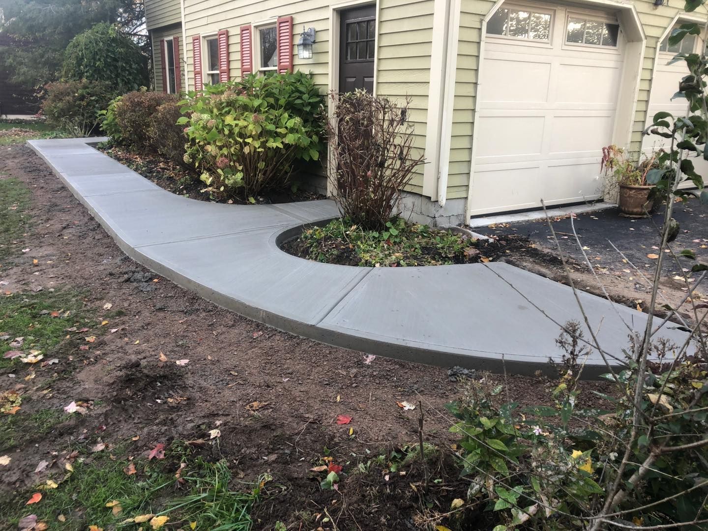 A new gray concrete walkway curves past a house with beige siding, a garage, and landscaping.