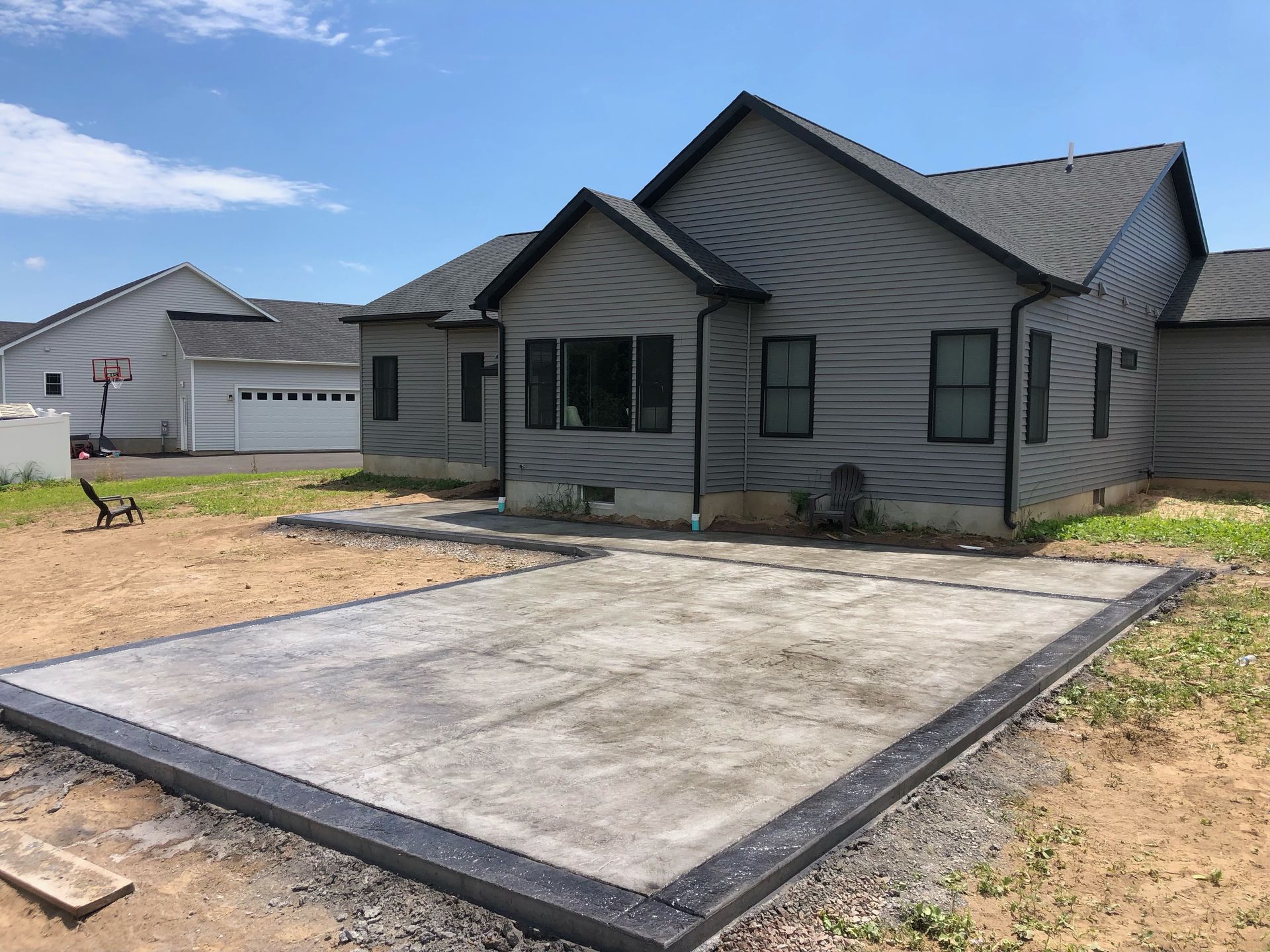 A house with gray siding and a concrete patio bordered by dark edging, on a sunny day.