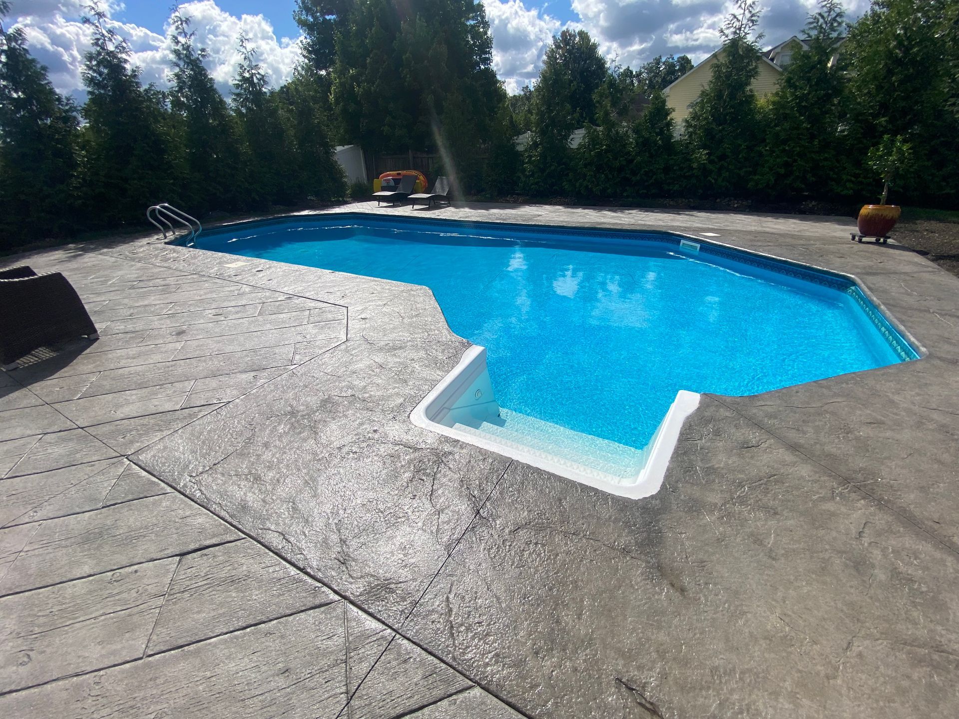 Blue pool surrounded by grey concrete patio, green trees, and blue sky.