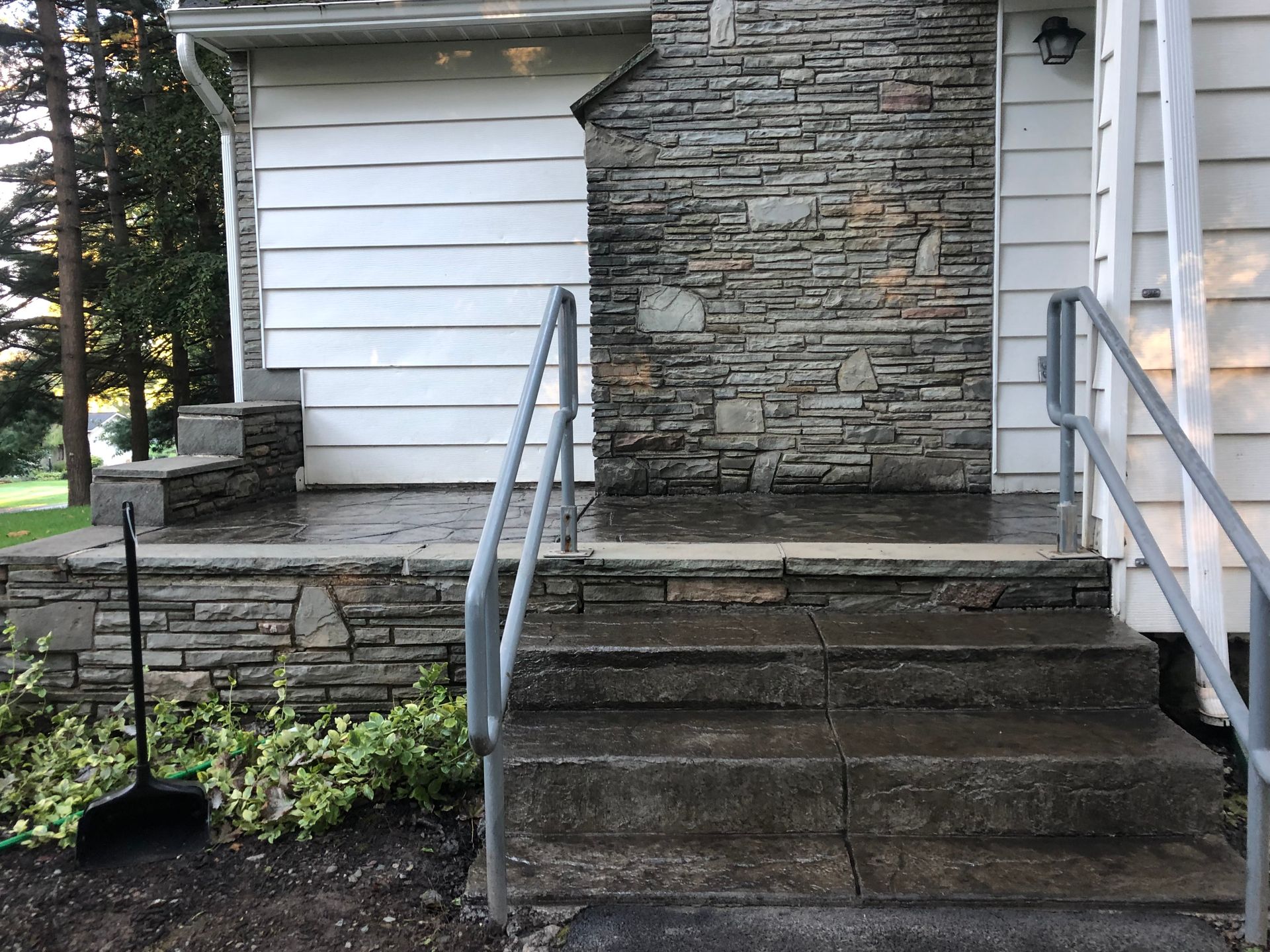 Stone steps leading to a house entrance with a stone wall, white siding, and metal handrails.