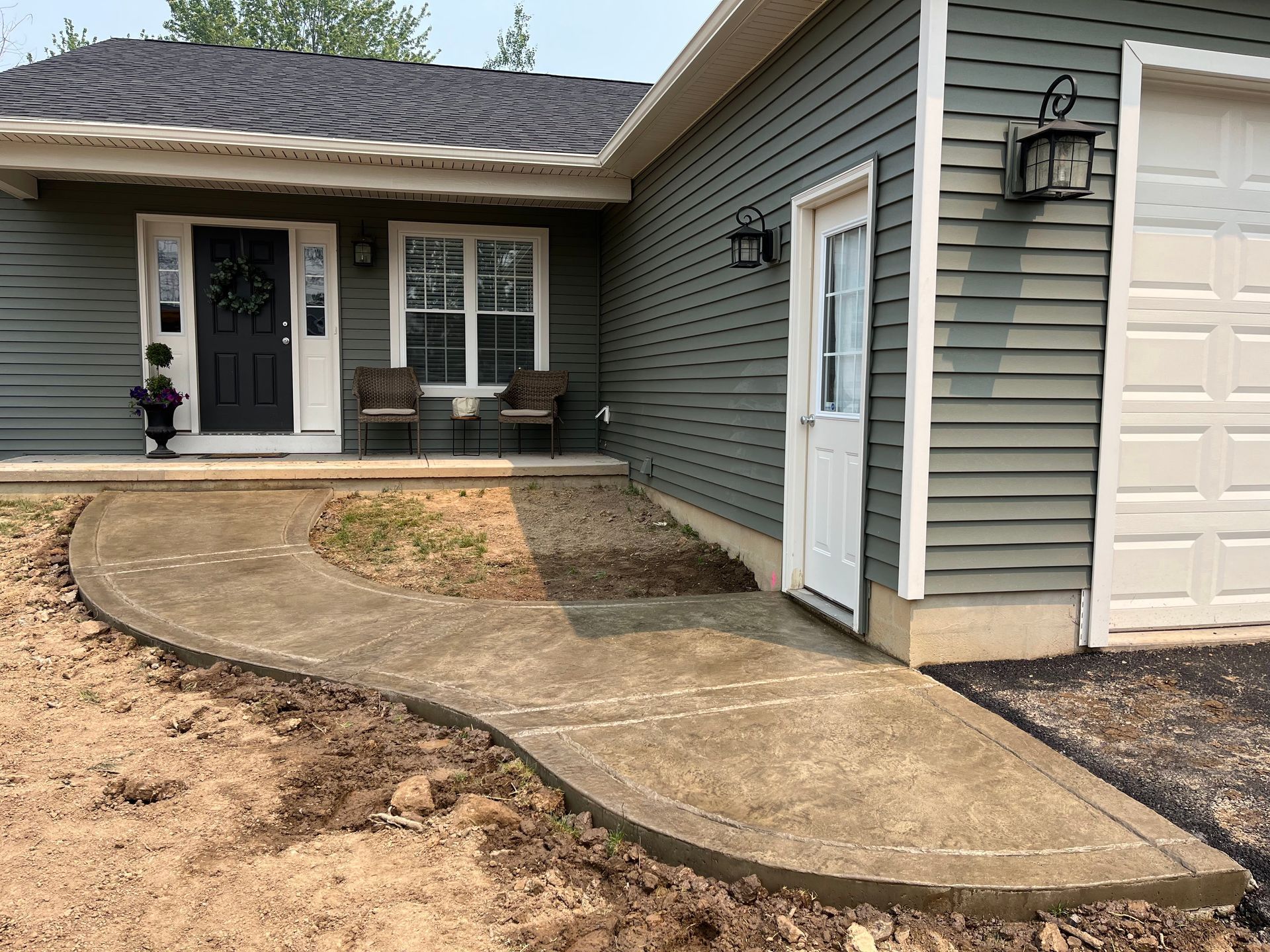 Concrete walkway leading to a green-sided house with black door and white garage door.