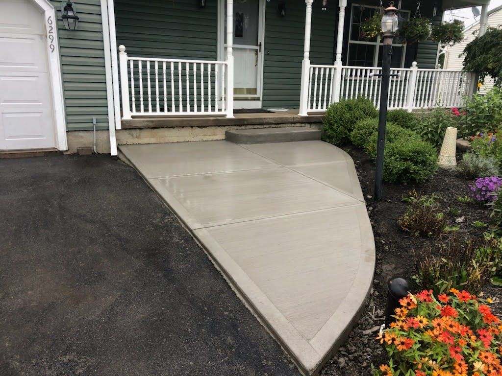 Newly poured concrete walkway leading to a house entrance with white railing and colorful flowers.