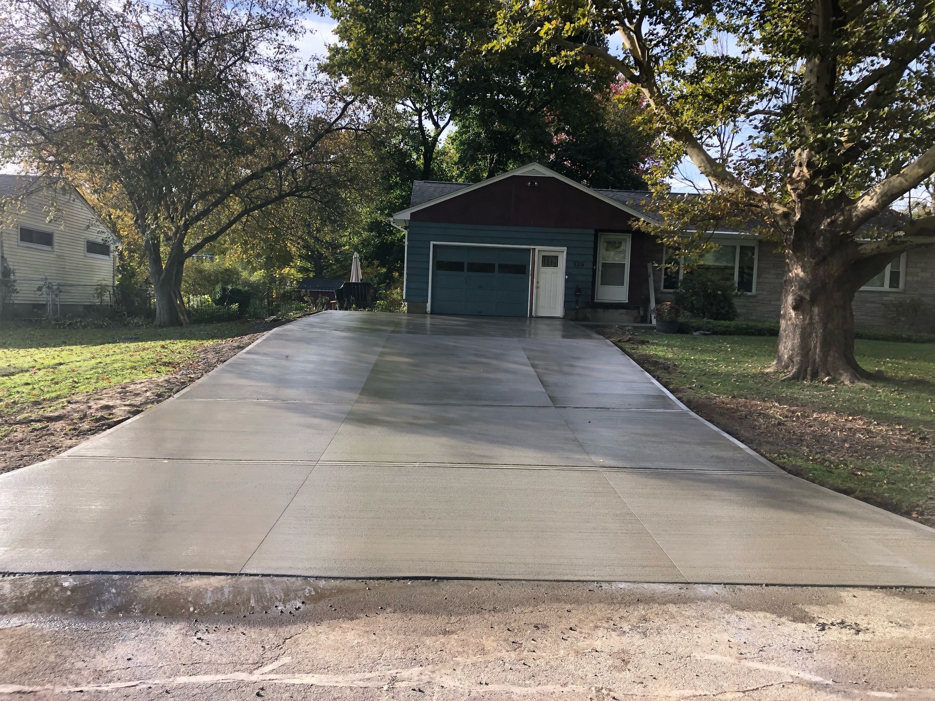 Newly poured concrete driveway leading to a one-story house with a green garage door and trees.