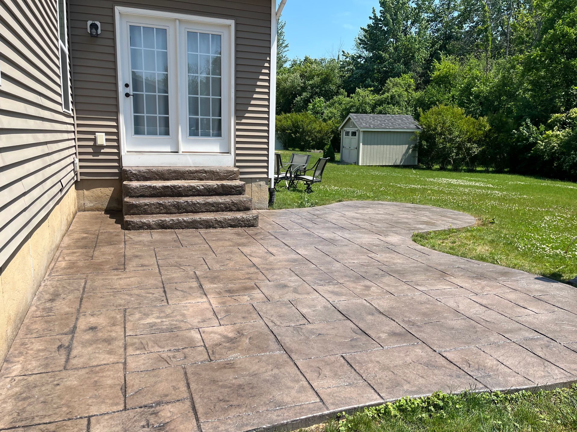A stone patio with steps leading to a house's double doors, with a shed and green lawn in the background.