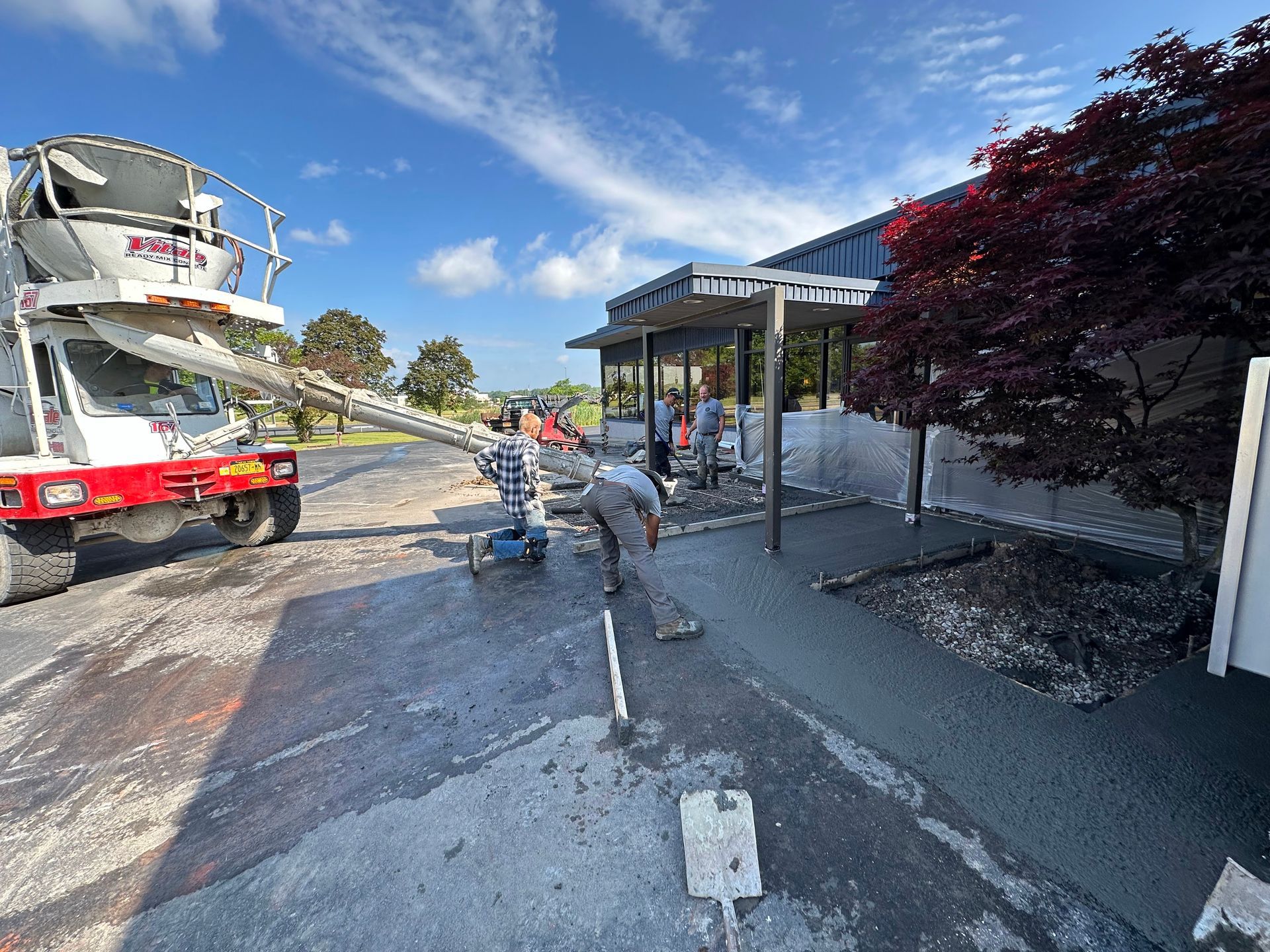 Concrete truck pouring cement for sidewalk construction at a building with workers on a sunny day.