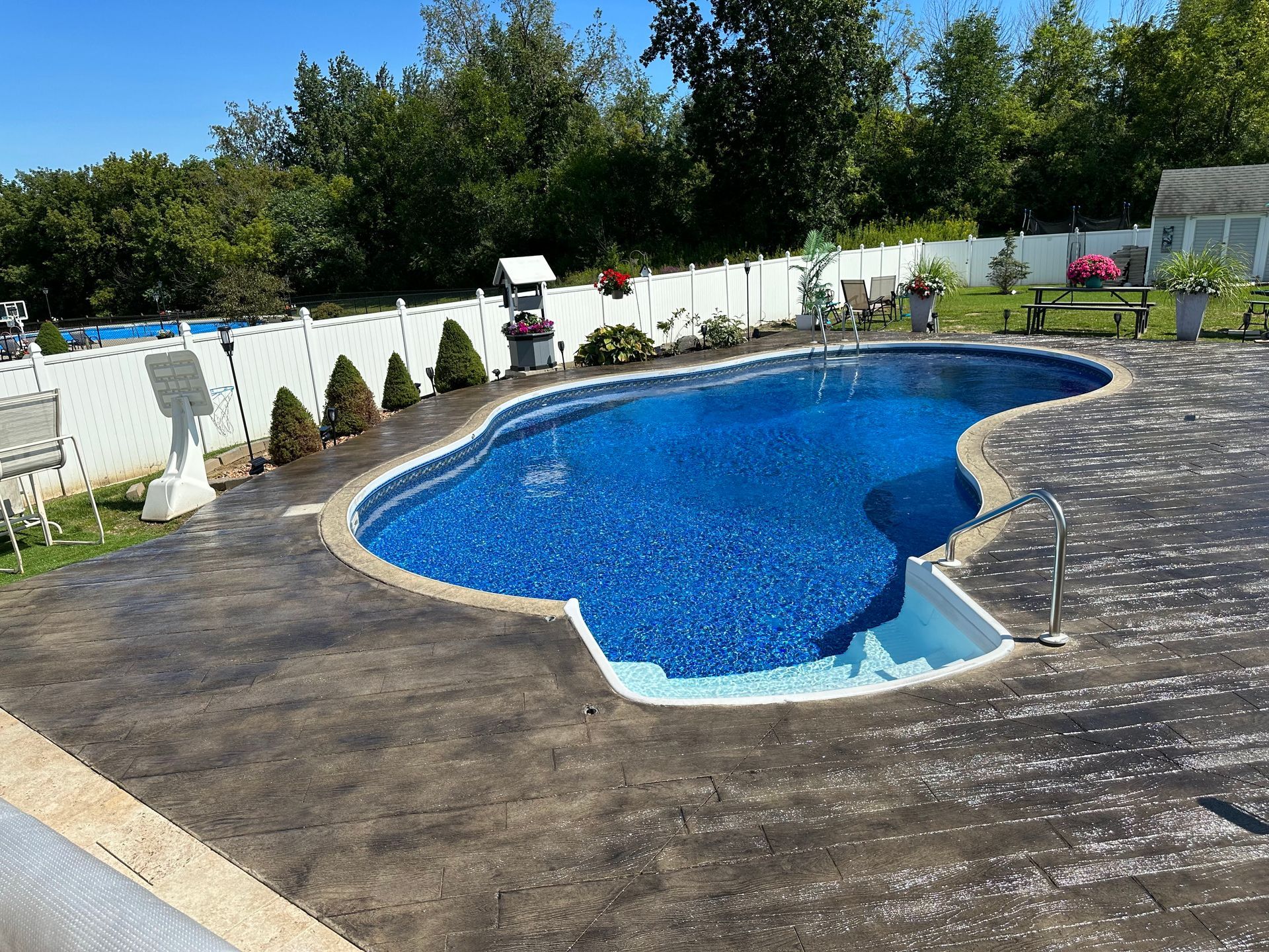 Swimming pool with blue water, surrounded by gray concrete and white fence, under a sunny sky.