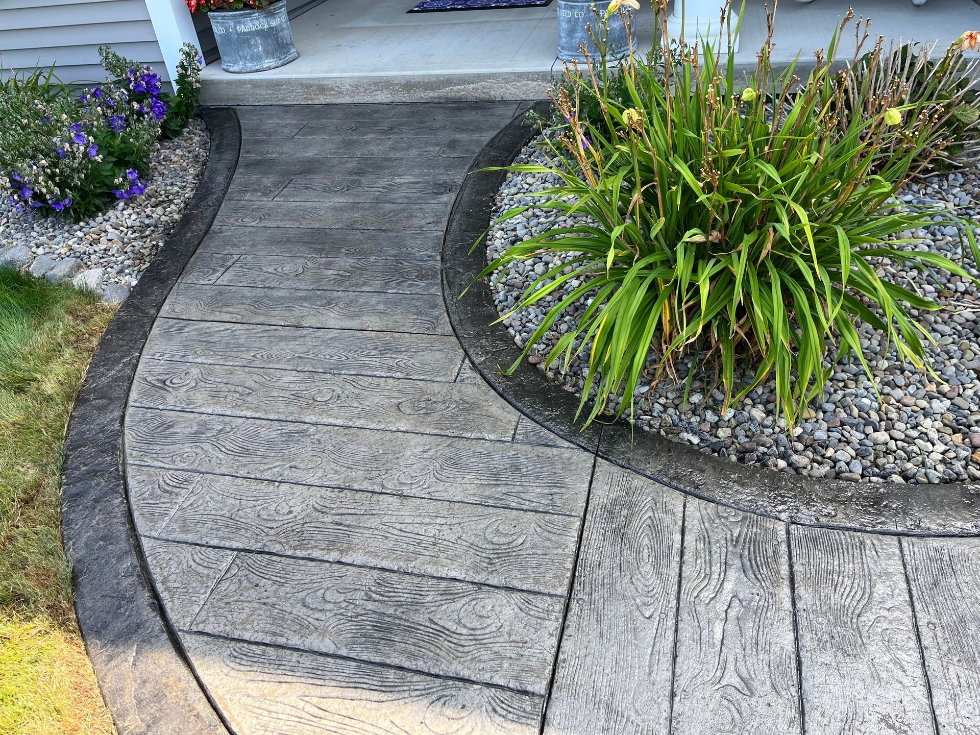 Curving concrete walkway with a wood grain texture, bordered by dark grey edges and landscaping.