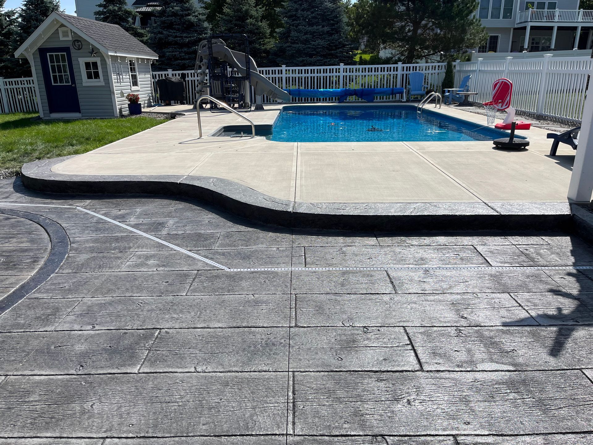 Concrete patio with a pool, small shed, and white fence in a sunny backyard.