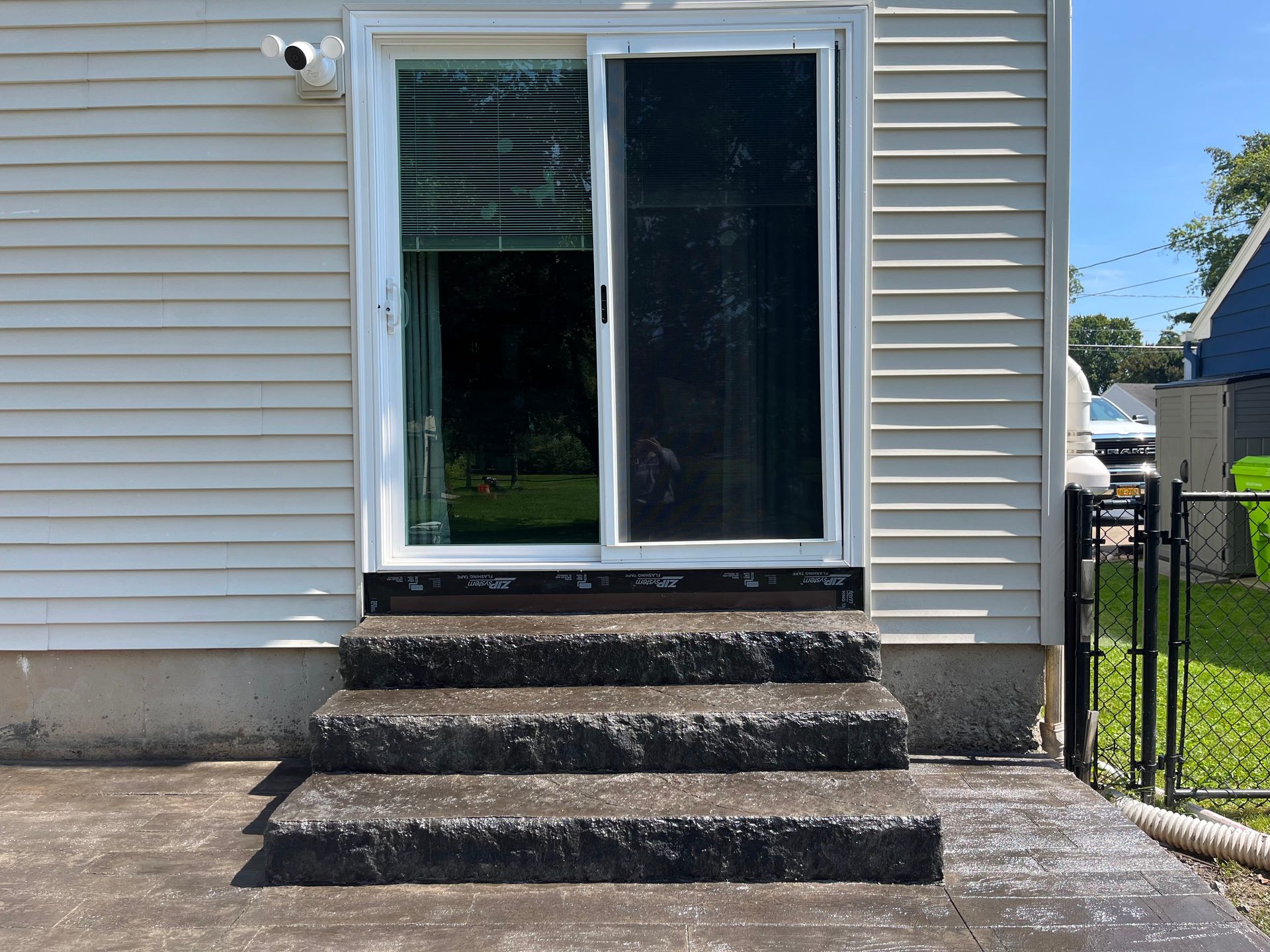 Sliding glass door with three concrete steps leading up. Exterior of a house on a sunny day.