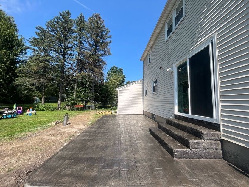 Backyard patio with concrete steps leading to sliding door of a two-story house; shed and trees in background.