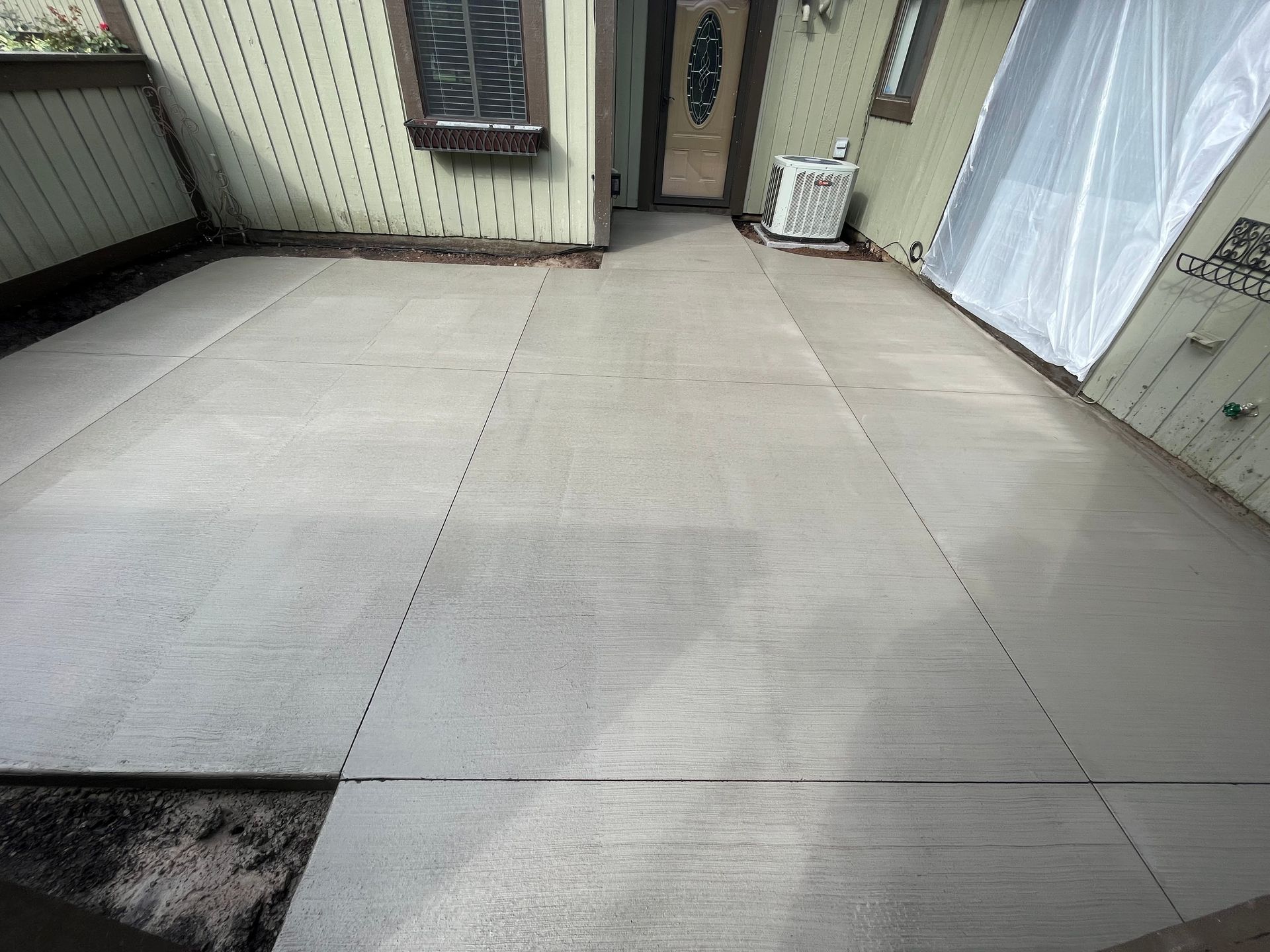 Newly poured concrete patio, light gray, with control joints, beside house, with door and air conditioner.