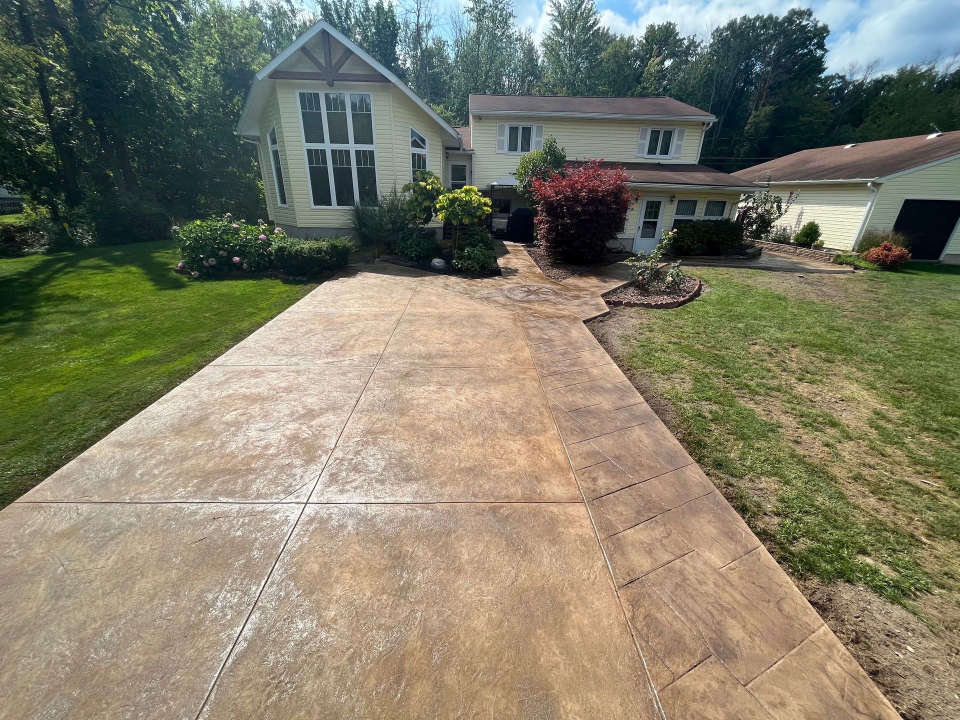 Concrete driveway leads to a yellow house with tall windows, surrounded by green grass and trees on a sunny day.