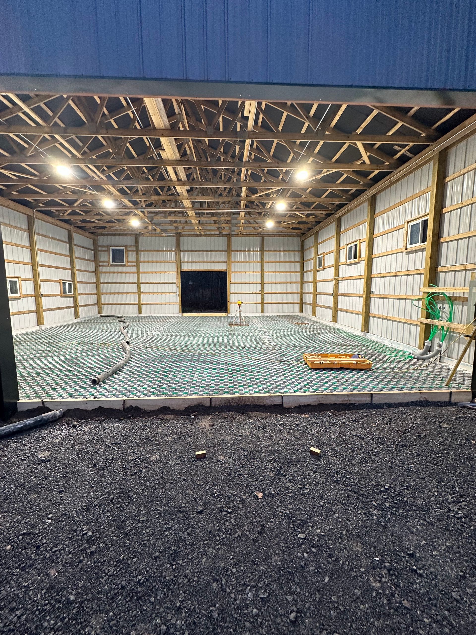 Interior of a building under construction, showing a grid of green pipes on a concrete floor. Walls and ceiling are exposed beams.