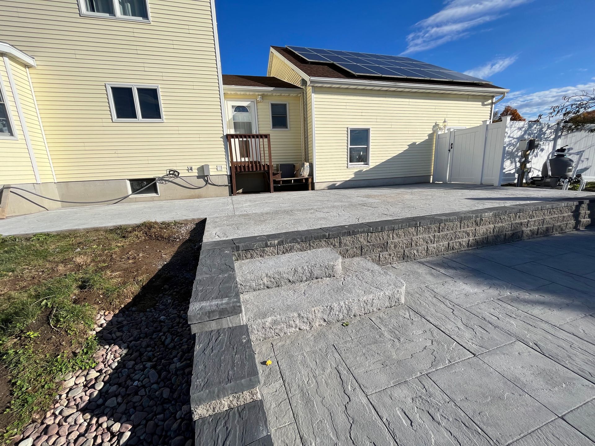 Backyard patio with stone steps and retaining wall, adjacent to a yellow house with solar panels.